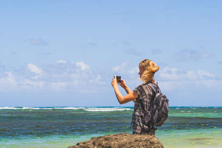 Woman takes picture on beach with smartphone against teal waters and clouds