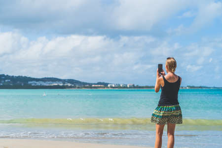 Woman using smartphone on beach to take pictures against teal waters and clouds