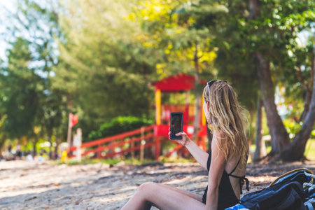 Woman takes selfie on beach with smartphone against teal waters and cloudsの写真素材