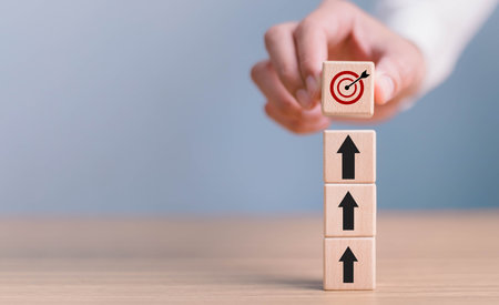 Business development to success and growing growth concept. Arrow leading point to target icon on wooden blocks with businessman hand placing red goal dartboard symbol.の写真素材