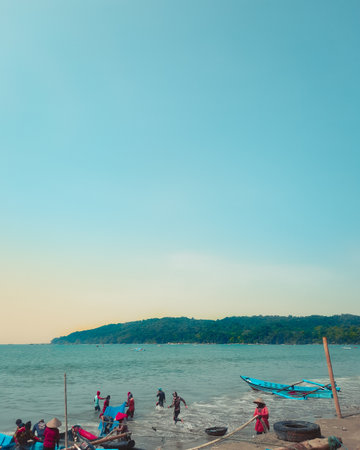 A calm beach scene where a group of people stands near the shoreline, facing gentle waves under a wide blue sky, with rocks and soft evening light creating a peaceful atmosphere.の写真素材