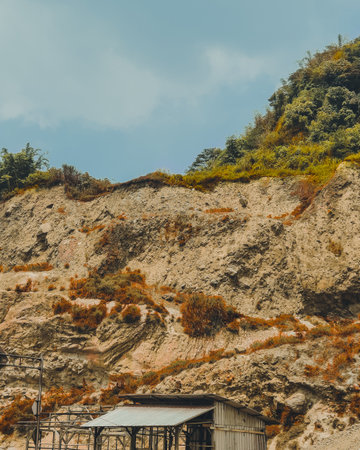 A rugged hillside with exposed rocky soil sits beneath a dense layer of green forest, creating a striking contrast between natural vegetation and eroded terrain under a clear blueの写真素材