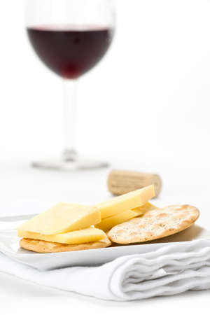 Cheese and crackers on a white appetizer plate with a glass of red wine and cork in the background against white.の写真素材