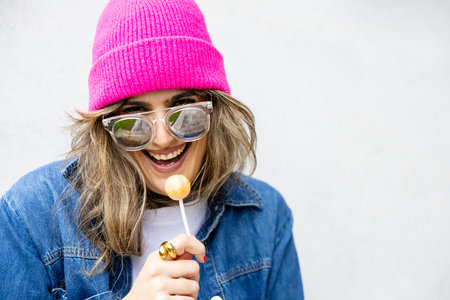 Portrait of woman with glasses and hat laughing cheerfully enjoying a lollipop, white spaceの写真素材