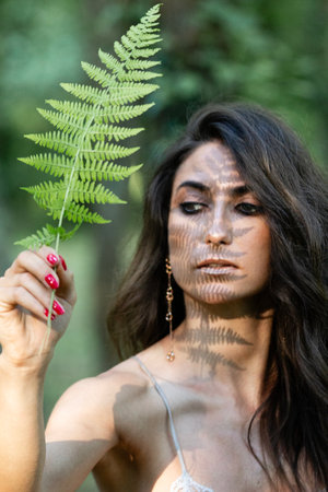 Portrait of brunette woman with fern leaf on her face in forest