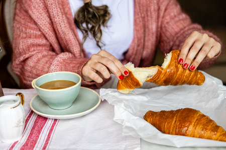 A woman in a cozy sweater tearing a croissant apart with coffee on the side, evoking a relaxed breakfast mood.の写真素材