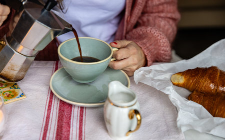 Close-up of hands pouring hot espresso from a mocha pot into a cup, with a delicious croissant awaiting.の写真素材