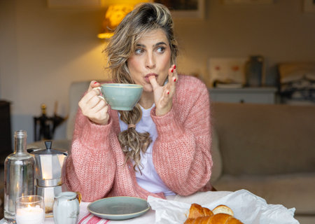 A young woman in a knit sweater appears pensive as she enjoys a cup of coffee, with pastries laid out in front.の写真素材