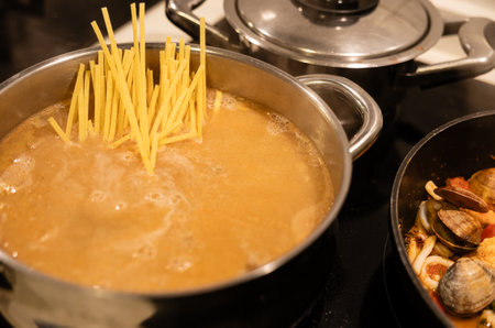 Golden pasta stands tall in boiling water next to a pot of simmering seafood stew on a stove.の写真素材