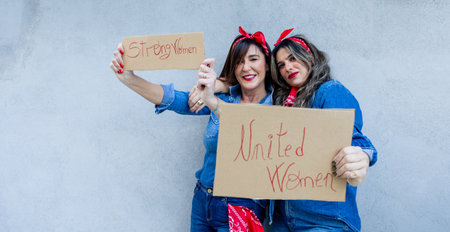Two enthusiastic women share a moment of solidarity, taking a selfie with signs 'Strong Women' and 'United Women', embodying empowerment and unity.の写真素材