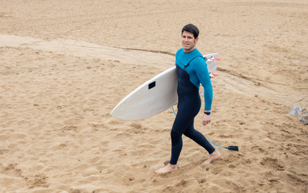 A young surfer in a blue and black wetsuit holding a surfboard, strolling on the sandy beach."の写真素材
