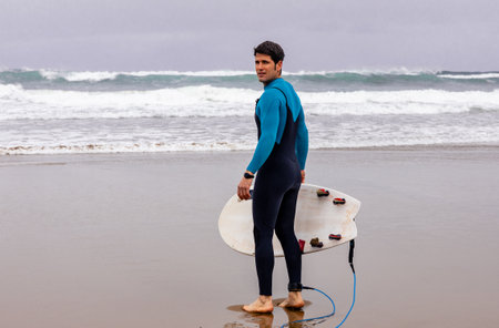 A man in a blue wetsuit with a white surfboard looks out at the ocean waves, contemplating the surf...