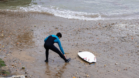 A surfer in a blue wetsuit secures his suit on a pebbly shore before a surf session, with his white surfboard beside him.の写真素材