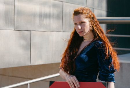 A red-haired woman in a velvet dress leans on a railing, her confident gaze enhancing the urban...