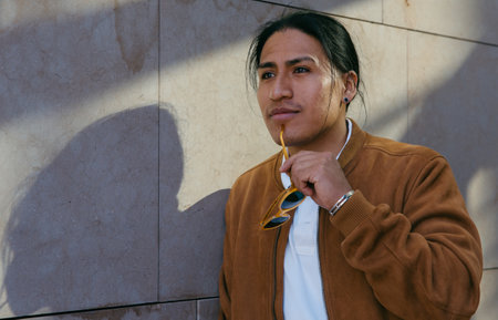 Young Ecuadorian man in a stylish brown jacket holding sunglasses, exploring the urban environment.