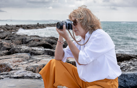 Elderly woman in a white shirt and orange pants sits on a rock by the sea, photographing the scenery...