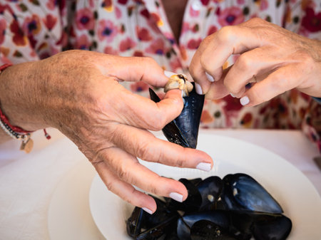 A close-up shot of hands skillfully opening a fresh mussel at a dining table. The image captures the details and textures of the seafood and the hands, set against a floral blouse backgroundの写真素材