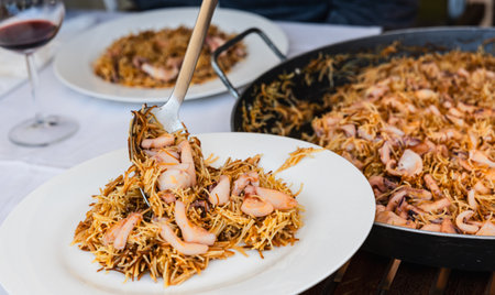 A close-up shot of a person serving seafood noodles from a large pan onto a plate at a dining table. The image captures the rich textures and inviting presentation of this traditional Mediterranean dish.の写真素材