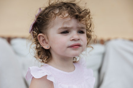 A close-up portrait of a two-year-old girl with curly hair, wearing a light pink dress and a bow.