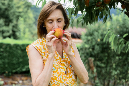 Mature woman savoring the scent of a ripe peach, standing in a lush garden. Capturing the essence of...