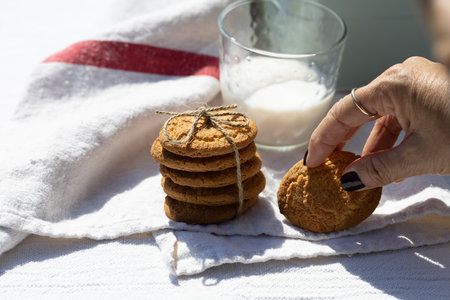 A hand reaches for a homemade cookie from a stack tied with twine, placed beside a glass of milk on...