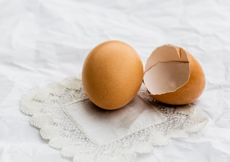 Close-up still life of a whole brown egg and a broken eggshell on a white fabric background.