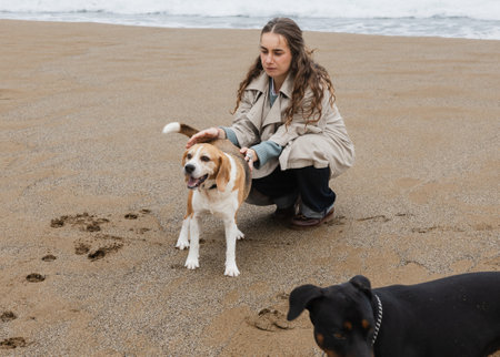 Woman crouching to pet her happy beagle on the beach by the ocean