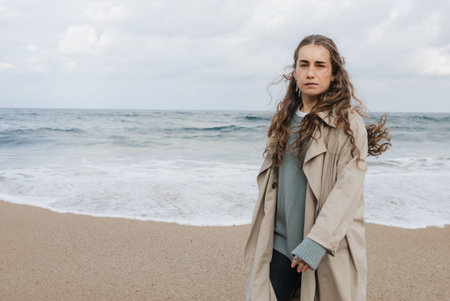 A young woman dressed in a trench coat stands along the beach shoreline.
