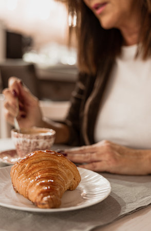 A golden croissant on a white plate with a blurred woman stirring coffee in the background.の写真素材