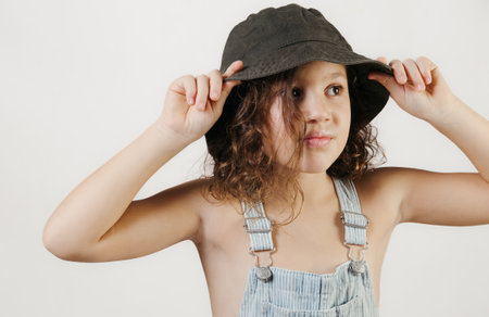Curly-haired girl in denim overalls posing with a black hat in studio.
