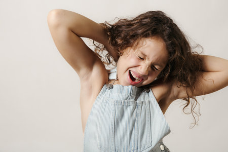 Girl with curly hair and denim overalls posing with a playful expression.の写真素材