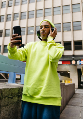 Young man in neon outfit using phone and headphones on city streetの写真素材