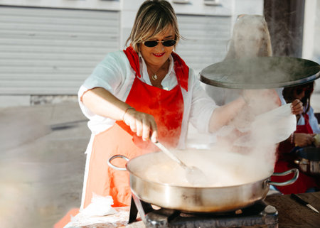 Caucasian middle-aged woman wearing a red apron stirs a steaming stew in a large metal pot at a festive outdoor community cooking event.の写真素材