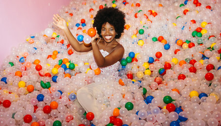 Young Black woman having fun in a colorful ball pool while smiling at the camera.