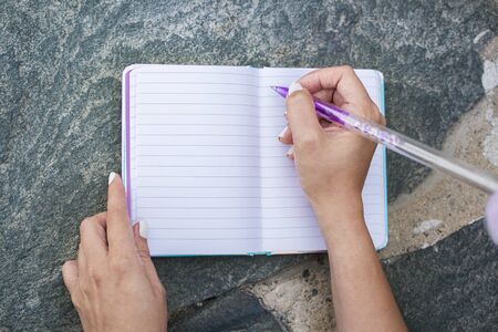 Woman's hands writing in her personal diary, on a rock, outdoors.