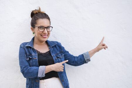 Smiling woman with glasses and denim jacket isolated on white background, pointing to the right with...
