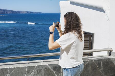 Woman taking photos with her smartphone to a summer landscape with sea and white houses lit by the sun.の写真素材