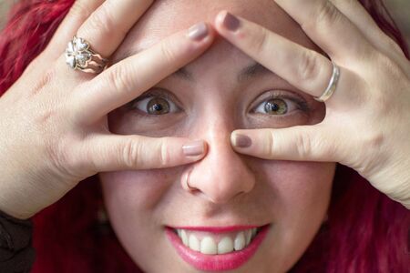 Close up portrait of a girl with green eyes and reddish hair covering her face with her hands revealing her eyes and her huge smile.の写真素材