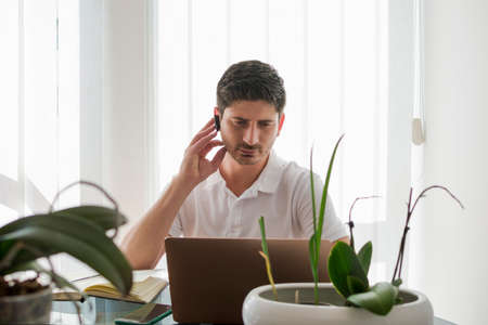 Young man working in his work zone, talking with an earpiece in his ear and surrounded by plants.