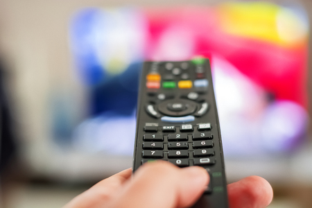 A hand pressing a remote control button to change channels in front of a blurry television screenの写真素材