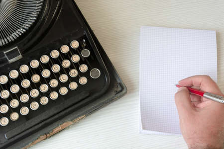a male hand writing on a white checkered paper with a red pen next to an old wrecked black...