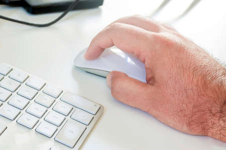 the white hand of a caucasian man working on a computer clicking a white mouse near a computer...