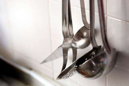 A skimmer, two forks and two metal ladles hanging on the tiled kitchen wall. Metal kitchen utensilsの写真素材