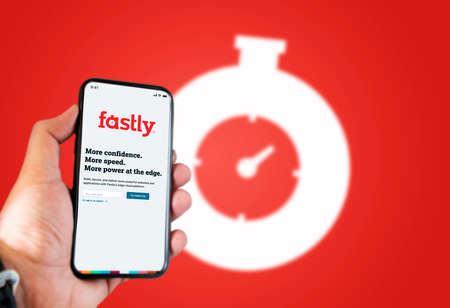 San Francisco, CA, USA, June 2021: A hand holding a phone with the Fastly app on the screen. Red background with blurred Fastly logo. Fastly is an American cloud computing services provider.のeditorial素材