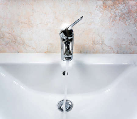 Chromed faucet with lever to mix hot and cold water on a modern white sink in a bathroom with tiled walls. Domestic life and hygieneの写真素材