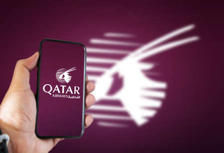 Doha, Qatar, July 2021: A hand holding a phone with the Qatar Airways airline app on the screen and the arabian oryx logo blurred on a dark cherry background.のeditorial素材