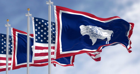 Wyoming and American flags waving under blue sky with bison emblem. Concept of patriotism, state...