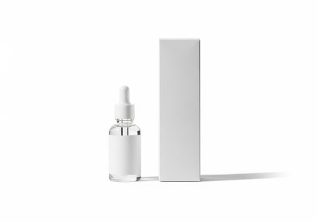 A clear glass dropper bottle with a silver cap stands next to a tall, slim white cardboard box on a clean white surface.の素材