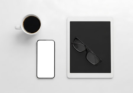 An overhead shot of a clean and minimalist workspace featuring a smartphone with a blank screen, a tablet displaying glasses, and a hot cup of coffee, perfect for remote work or study setups.の素材