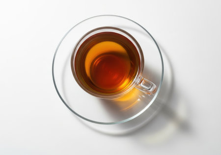 A top-down shot of a transparent glass cup holding dark amber tea, resting on a clear glass saucer against a white background.の素材